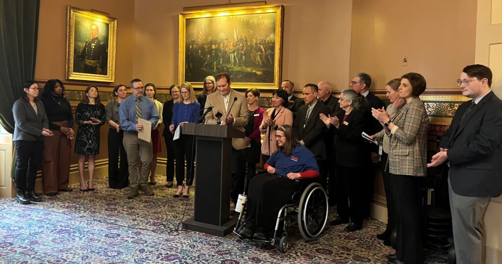 Nord and advocates gather around a microphone at an indoor press conference in the vermont state capitol building calling for step therapy reform.